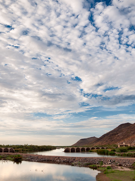 Fluffy clouds scatter across the sky above Gillespie Dam, mirroring on the water's surface in a tranquil scene in Arlington, Arizona.