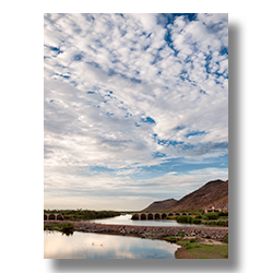 Fluffy clouds scatter across the sky above Gillespie Dam, mirroring on the water's surface in a tranquil scene in Arlington, Arizona.