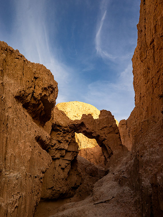 Shaded mudstone arch in a slot canyon, dramatically backlit by afternoon sun, under a sky with wispy cirrus clouds near Alamo Lake, Arizona.