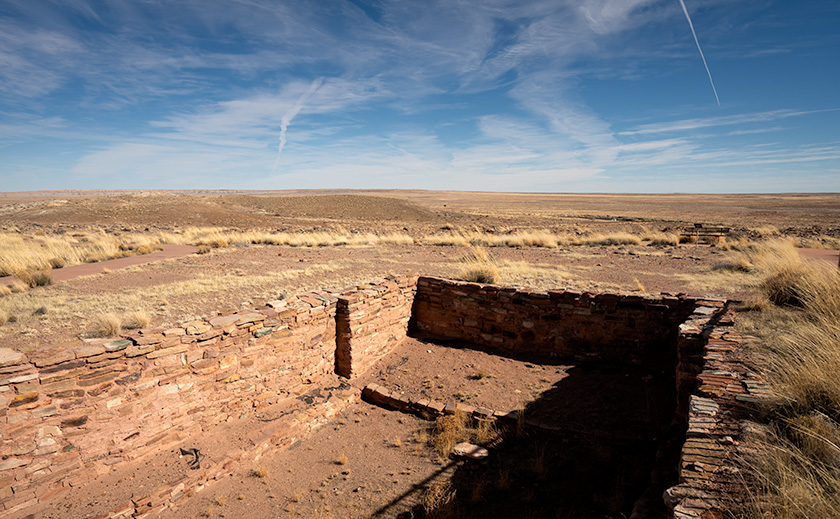 Ruins of a kiva at Homolovi State Park under a vast sky marked by crossing contrails.