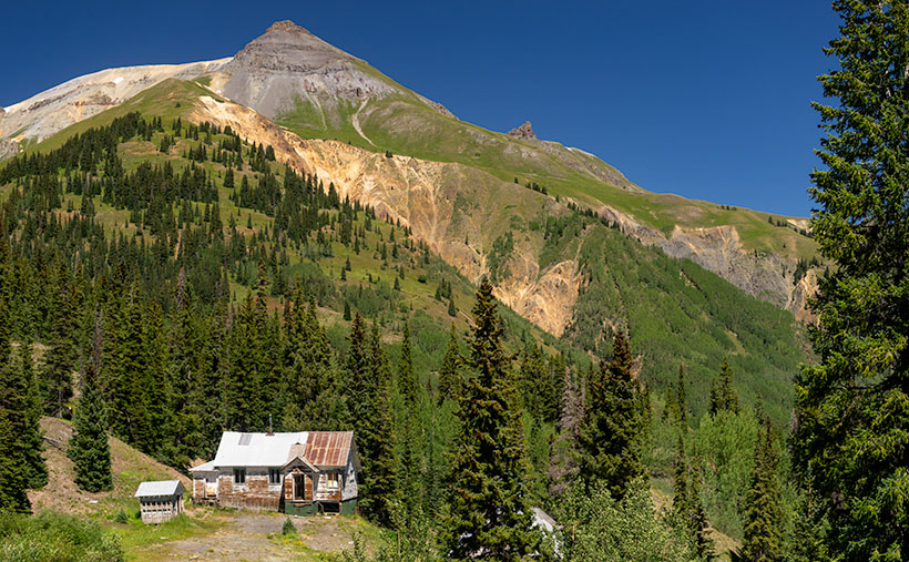 Rustic cabin nestled at the base of towering mountains with lush greenery and a clear blue sky.
