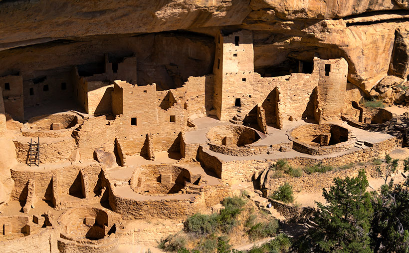 Sunlit kivas in front of multi-level ancient dwellings at Sunset House, framed by an arching, varnished cave wall with trees in the lower right corner.