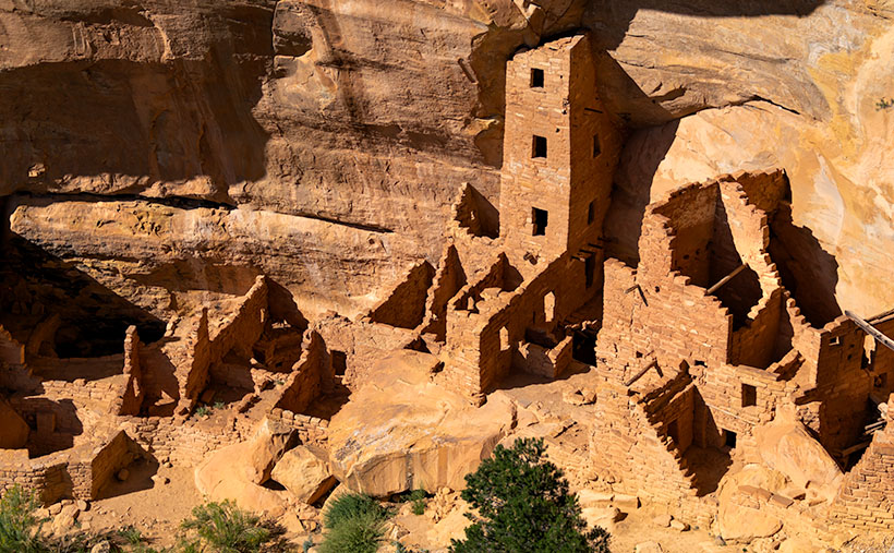 Aerial view of Tower House ruins in Mesa Verde, capturing a four-story tower and adjacent apartment-like structure against a backdrop of varnished walls.