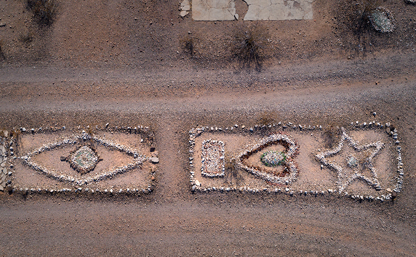 Drone image of two stone glyphs in Sundad, Arizona: a left-pointing arrow and a diamond-shaped eye with blue glass center, split by a cracked concrete slab