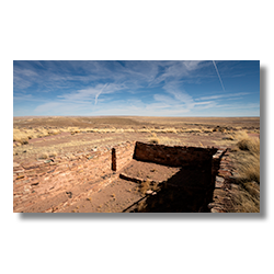 Ruins of a kiva at Homolovi State Park under a vast sky marked by crossing contrails.