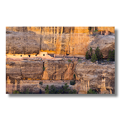 A layered sandstone canyon wall streaked with black varnish, housing light-colored ancient ruins.