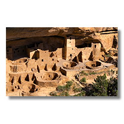 Sunlit kivas in front of multi-level ancient dwellings at Sunset House, framed by an arching, varnished cave wall with trees in the lower right corner.