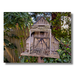 Old wooden barn-shaped birdhouse with broken boards, nestled among garden plants at Cambria Pines Lodge.