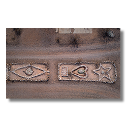 Drone image of two stone glyphs in Sundad, Arizona: a left-pointing arrow and a diamond-shaped eye with blue glass center, split by a cracked concrete slab