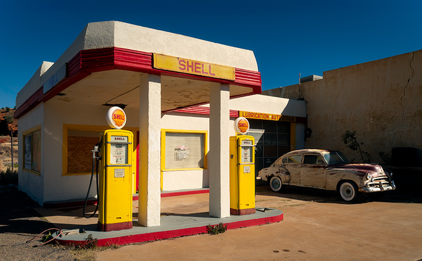 A retro Shell gas station in Layton, Arizona, with bright yellow pumps and a classic car parked nearby, framed by blue skies and mid-century architecture.